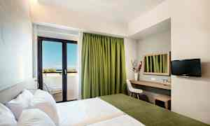 Ett hotellrum med två sängar, en arbetsyta med spegel, en stol och en TV på väggen. Rummet har en balkong med gröna gardiner och utsikt mot havet. (AI-genererad text)