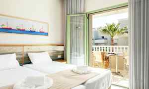 Ett ljust hotellrum med två enkelsängar, vita sängkläder och beige överkast. Ett inslag av båtmotiv hänger på väggen ovanför sängen. Rummet har en öppen balkongdörr med utsikt över en palm och ett bord med två stolar. (AI-genererad text)