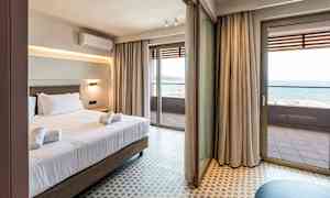 Hotellrum med säng och handdukar, stora glasdörrar som leder ut till en balkong som visar havet. (AI-genererad text)