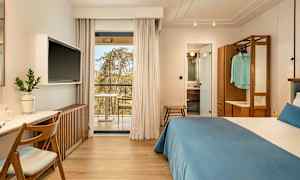 Hotellrum med dubbelsäng, skrivbord och TV. Glasdörrar leder ut till en balkong. Badrum synligt genom en öppen dörr. (AI-genererad text)