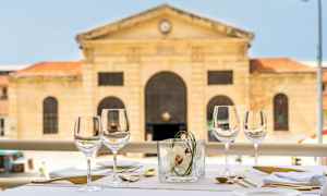 Ett dukat bord utomhus med vinglas, bestick och servetter. I bakgrunden syns en historisk stenbyggnad. (AI-genererad text)