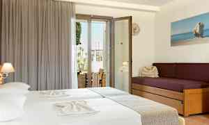 Ett hotellrum med två sängar klädda i ljusa lakan, en lila soffa med kuddar, och en glasdörr som leder ut till en balkong. (AI-genererad text)