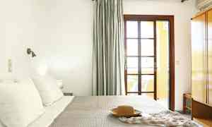 Ett sovrum med en säng, kuddar och en filt. Ett fönster med gardiner och ett nattduksbord syns. (AI-genererad text)