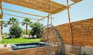 Bild på en uteplats med jacuzzi, hängstol och trämöbler, omgiven av träd och buskar. (AI-genererad text)