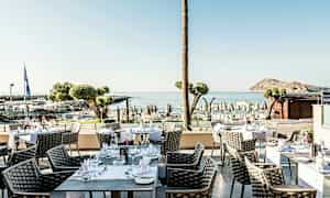 Uteservering med bord och stolar, havsutsikt och strand i bakgrunden. (AI-genererad text)