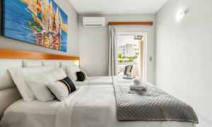 Dubbelrum med vita väggar, en dubbelsäng med grå överkast och en tavla på segelbåtar på väggen. (AI-genererad text)