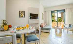 Ett ljust hotellrum med matbord och bakverk på en tallrik, soffgrupp och en balkong i bakgrunden. (AI-genererad text)