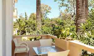 En balkong med bord och stolar, omgiven av gröna växter och palmer. (AI-genererad text)