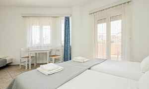 Ett ljust rum med två enkelsängar, ett bord med två stolar och fönster täckta av vita gardiner. (AI-genererad text)