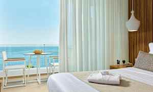 Utsikt från ett rum med en bäddad säng mot en balkong med stolar, bord och havsutsikt. (AI-genererad text)