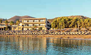 En semesteranläggning vid stranden med solstolar och parasoller i förgrunden och ett hotell bakom. (AI-genererad text)