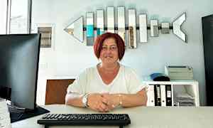 Person sitter vid ett skrivbord med tangentbord, datorskärm och hyllor i bakgrunden. (AI-genererad text)