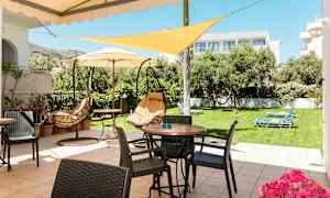 En terrass med bord, stolar och hängstolar, omgiven av en gräsmatta med solstolar och träd. (AI-genererad text)