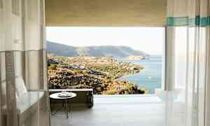 Utsikten från ett rum med gardiner mot ett bergigt landskap vid kusten och havet. (AI-genererad text)