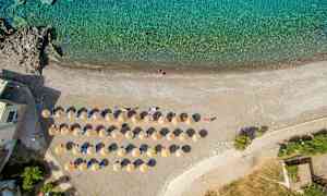 Flygbild över en strand med uppställda solstolar och parasoller vid kanten av turkost vatten. (AI-genererad text)