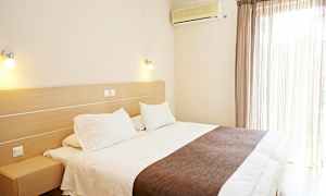 Ett hotellrum med en stor säng, vita sängkläder och brunt överkast. En luftkonditionering är monterad på väggen bredvid fönstret med ljusa gardiner. (AI-genererad text)