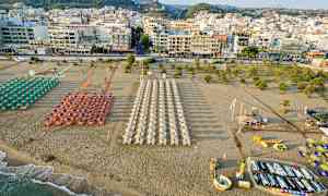En strand med parasoller och solstolar uppställda i rader framför en stad med många byggnader. (AI-genererad text)