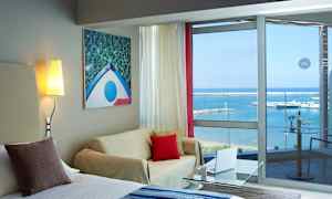 Ett hotellrum med sittgrupp, ljusmålning på väggen och glasfönster med utsikt över havet och en småbåtshamn. (AI-genererad text)