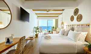 Ett hotellrum med vita väggar, en bäddad säng, sittmöbler och en balkong med utsikt över havet. (AI-genererad text)