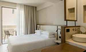 Ett hotellrumsinteriör med en dubbelsäng, ett fönster med gardiner, en balkong med stol och ett tvättställ. (AI-genererad text)