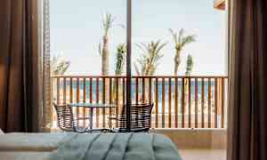 Hotellrum med utsikt över en terrass med bord och stolar, följt av palmer och havet i bakgrunden. (AI-genererad text)