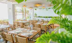 En restaurang med bord och stolar i trä, omgiven av grönska och med utsikt över havet. (AI-genererad text)