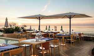 En utomhusrestaurang vid havet under solnedgången med bord och stolar samt stora parasoll. (AI-genererad text)