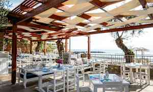 Uteservering med vita bord och stolar under ett tak av trä. Havsutsikt i bakgrunden. (AI-genererad text)