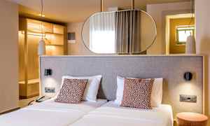 Ett hotellrum med två enkelsängar, dekorativa kuddar och en modern inredning. Bakom sängen finns en spegel och garderob i trä. (AI-genererad text)