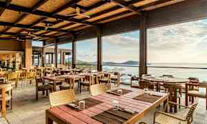 En öppen och ljus restaurang med träbord och stolar, med utsikt över havet och horisonten. (AI-genererad text)