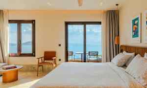 Ett hotellrum med en bäddad säng, stolar och ett bord vid fönster med utsikt mot havet. (AI-genererad text)