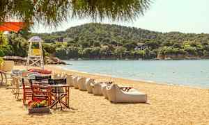 En sandstrand med solstolar och bord, med gröna kullar i bakgrunden. (AI-genererad text)