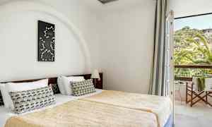 Rummet visar två enkelsängar med guldbeläckta täcken och en dekorativ kudde på varje säng. En väggprydnad hänger över huvudgaffeln, och ett fönster med utsikt över grönska och ett hus syns till höger. (AI-genererad text)