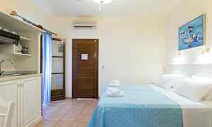 Ett hotellrum med två bäddar klädda i blå sängkläder, en träbeklädd dörr, ett mindre kök med diskho och en målning på väggen. (AI-genererad text)