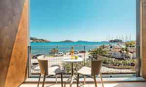 Utsikt från en balkong med två stolar och ett bord, mot havet och en småbåtshamn. (AI-genererad text)