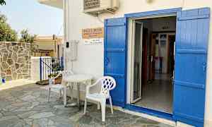 En vit byggnad med blå dörrar och fönsterluckor. Framför dörren står två vita plaststolar och ett litet bord. (AI-genererad text)