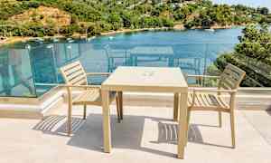 Två beige stolar och ett bord på en terrass med glasräcke och utsikt över en vik med båtar och grönska. (AI-genererad text)