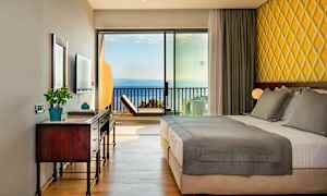 Hotellrum med dubbelsäng, skrivbord och havsutsikt genom glaspanel. (AI-genererad text)