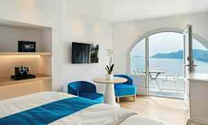 Hotellrum med ljusa vita väggar, möblerat med en säng, bord och stolar, och en balkong med utsikt över havet och en ö. (AI-genererad text)