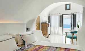 Ett ljust och modernt hotellrum med en säng, färgglad pläd, sittgrupp och stora fönster som vetter mot en utsikt över havet. (AI-genererad text)