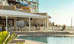 En utomhusrestaurang med vita möbler vid sidan av en pool, med några personer som sitter vid borden. Bakgrunden består av byggnader och en ljus himmel. (AI-genererad text)