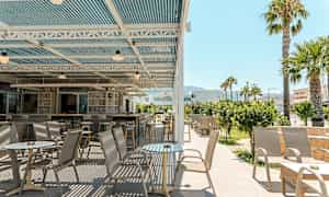 En utomhusterrass med bord och stolar under ett täckt tak, omgivande grönska och palmer. (AI-genererad text)