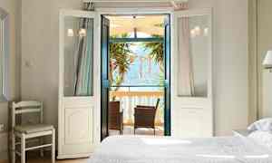 Ett ljust sovrum med öppna dörrar mot en balkong med möbler och utsikt över havet. (AI-genererad text)