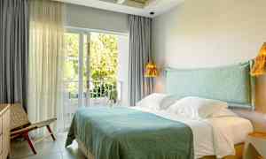 Hotellrum med dubbelsäng, blå sängkläder, och stora fönster som vetter mot en grönskande balkong. (AI-genererad text)