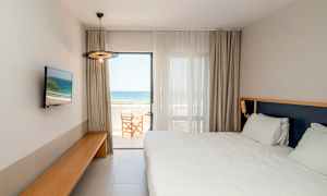 Ett hotellrum med vit säng, en TV på väggen och en balkong med havsutsikt. (AI-genererad text)