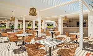 En utomhusrestaurang med sittplatser och bord, täckt med vita pergolastänger och dekorerad med rottinglampor. Baren är synlig i bakgrunden. (AI-genererad text)