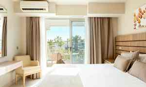 Ett hotellrum med en säng, ett skrivbord och en stol, samt en balkong med utsikt över palmer. (AI-genererad text)