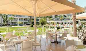 Utomhusrestaurang med möblerade bord nära en pool och omgivande byggnader. (AI-genererad text)