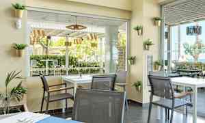 Ett modernt inrett café med bord och stolar, utsikt mot grönskande träd och utemöbler. (AI-genererad text)