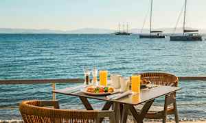 Ett bord med frukost och juice står vid havet, med segelbåtar i bakgrunden. (AI-genererad text)
